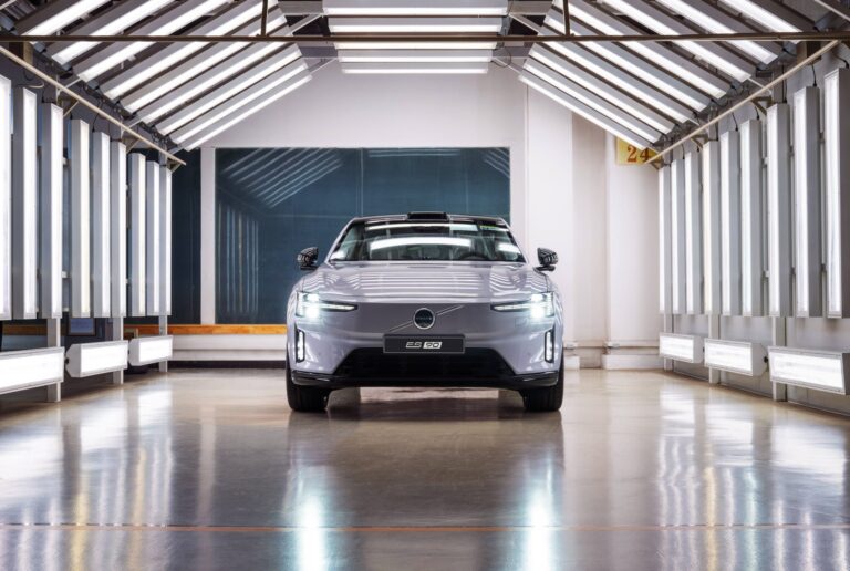 Widok z przodu na srebrne Volvo ES90 w jasno oświetlonym tunelu hali produkcyjnej.