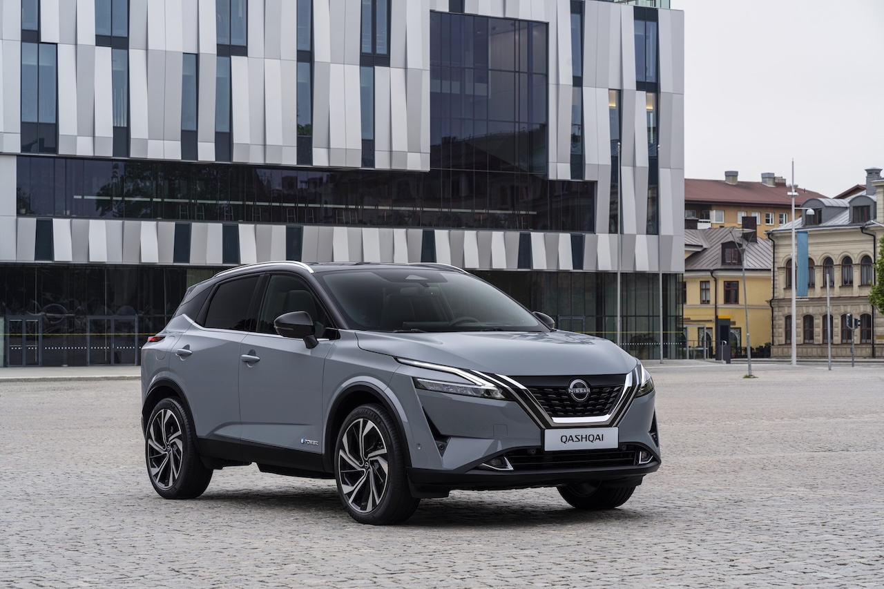 Szary SUV Nissan Qashqai zaparkowany na kamiennym placu na tle nowoczesnego budynku o nieregularnej, lśniącej, srebrno-szarej fasadzie geometrycznej.