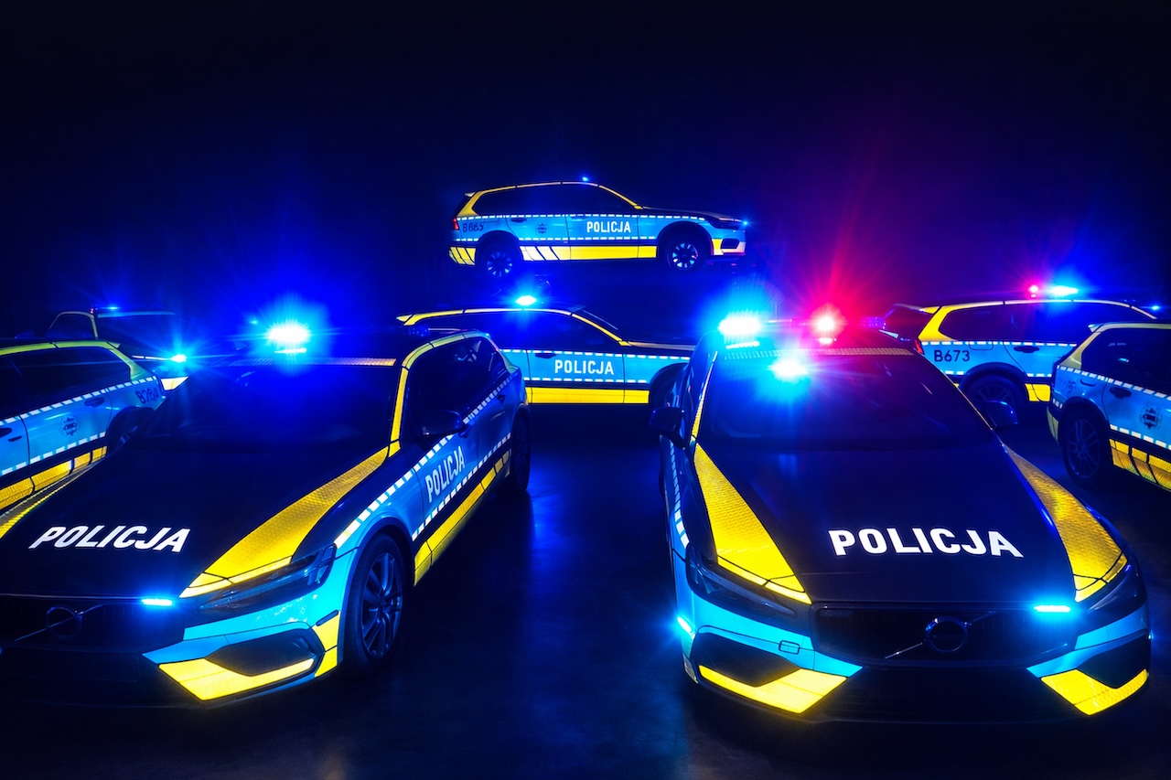 Kilka radiowozów Volvo z aktywnymi sygnałami błyskowymi w ciemnym pomieszczeniu, eksponujących odblaskowe napisy „POLICJA”.