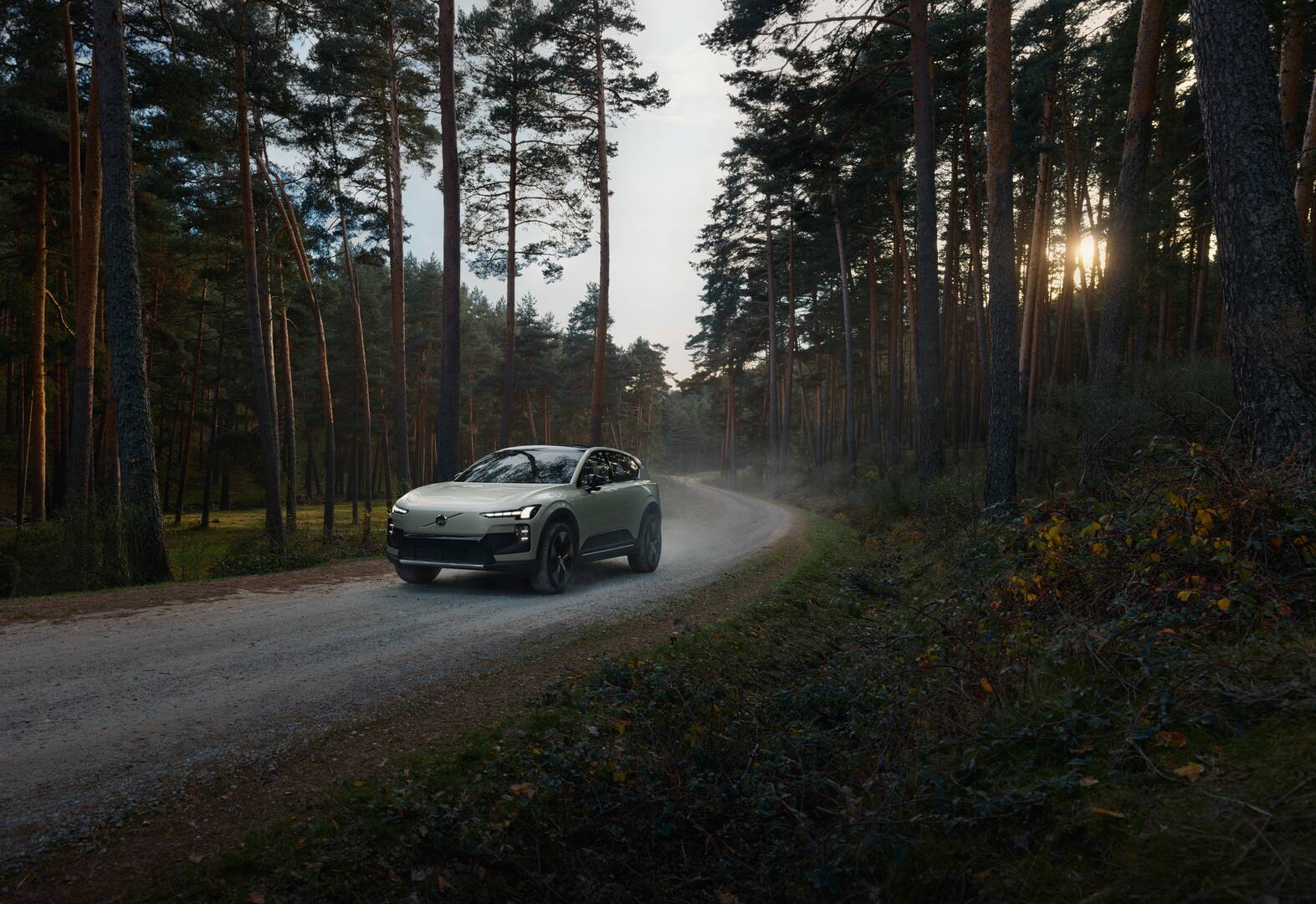 Volvo EX60 Cross Country jadące szutrową drogą przez las w świetle zachodzącego słońca.