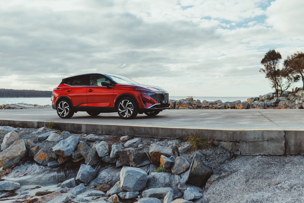 Czerwony SUV Nissan Qashqai zaparkowany na betonowej drodze tuż przy kamienistym wybrzeżu, w tle widoczny ocean oraz zachmurzone niebo.
