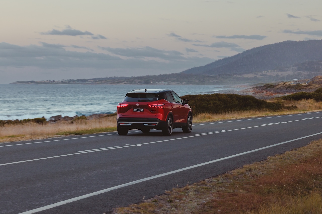Czerwony Nissan Qashqai sfotografowany od tyłu podczas jazdy asfaltową drogą wzdłuż oceanu, w tle widoczne pasmo górskie pod wieczornym niebem.