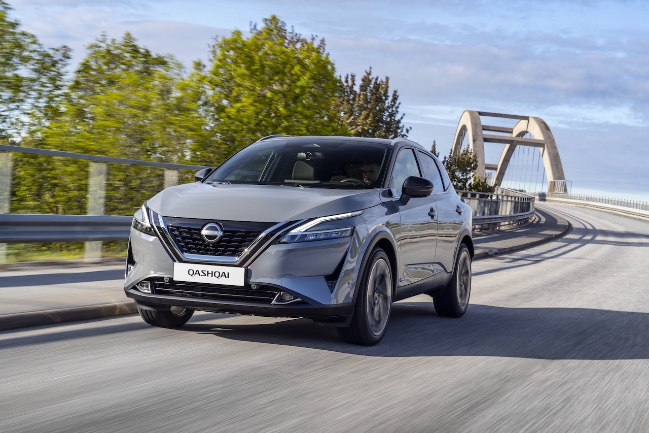Szary Nissan Qashqai w dynamicznym ruchu na drodze prowadzącej przez nowoczesny, betonowy most z łukową konstrukcją, w otoczeniu zielonych drzew.