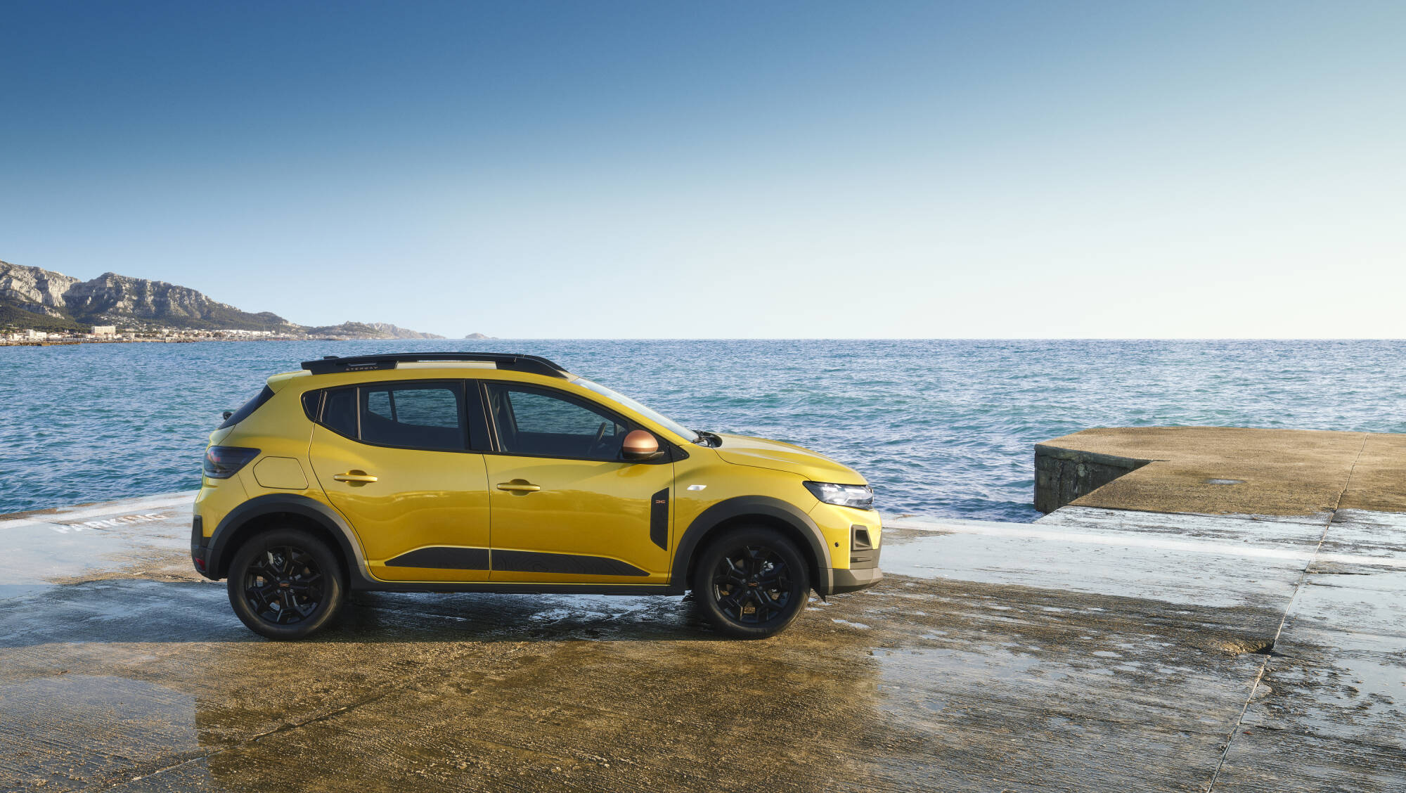 Profil Dacii Sandero w kolorze Sandstone zaparkowanej przy morzu, widoczne relingi dachowe i czarne felgi.