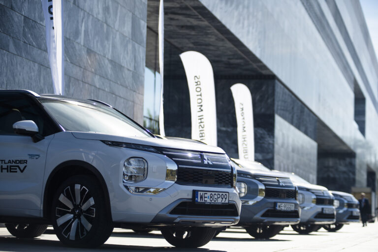 Szereg nowych modeli Mitsubishi Outlander PHEV zaparkowanych przed nowoczesnym budynkiem z szarą elewacją i białymi flagami Mitsubishi Motors.