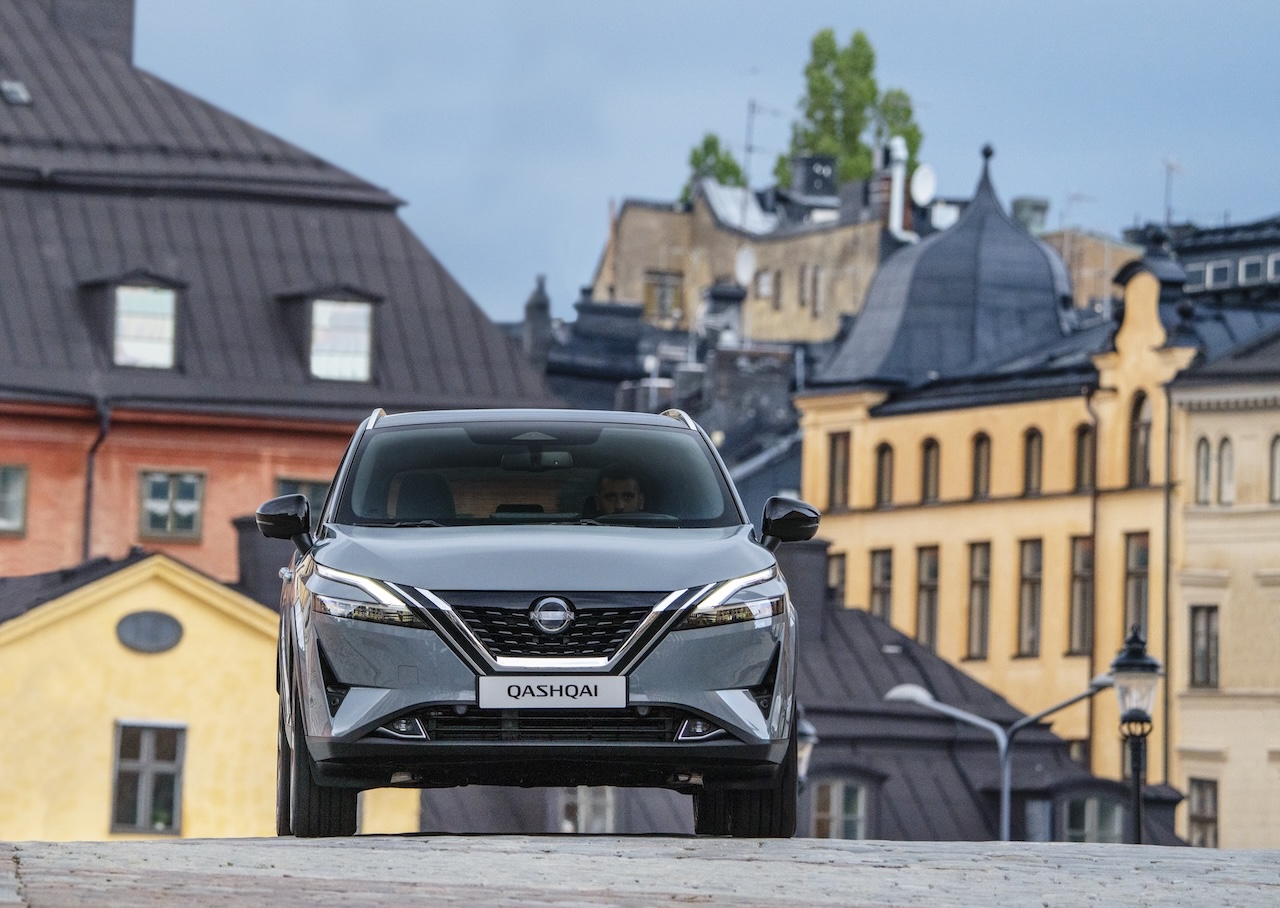 Szary Nissan Qashqai jadący w dół brukowanej ulicy miejskiej, w tle zabytkowe kamienice o żółtych elewacjach z ciemnymi dachami.