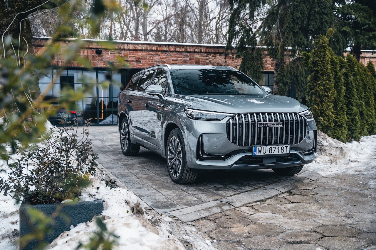 Szary SUV JAECOO 8 zaparkowany na podjeździe przed nowoczesnym budynkiem z cegły i szkła. Widoczny przedni grill z pionowymi chromowanymi listwami oraz logo marki.