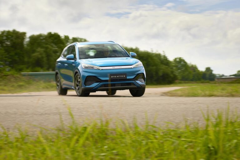 Niebieski SUV BYD ATTO 3 jadący po asfaltowej drodze w słoneczny dzień na tle zielonych drzew.