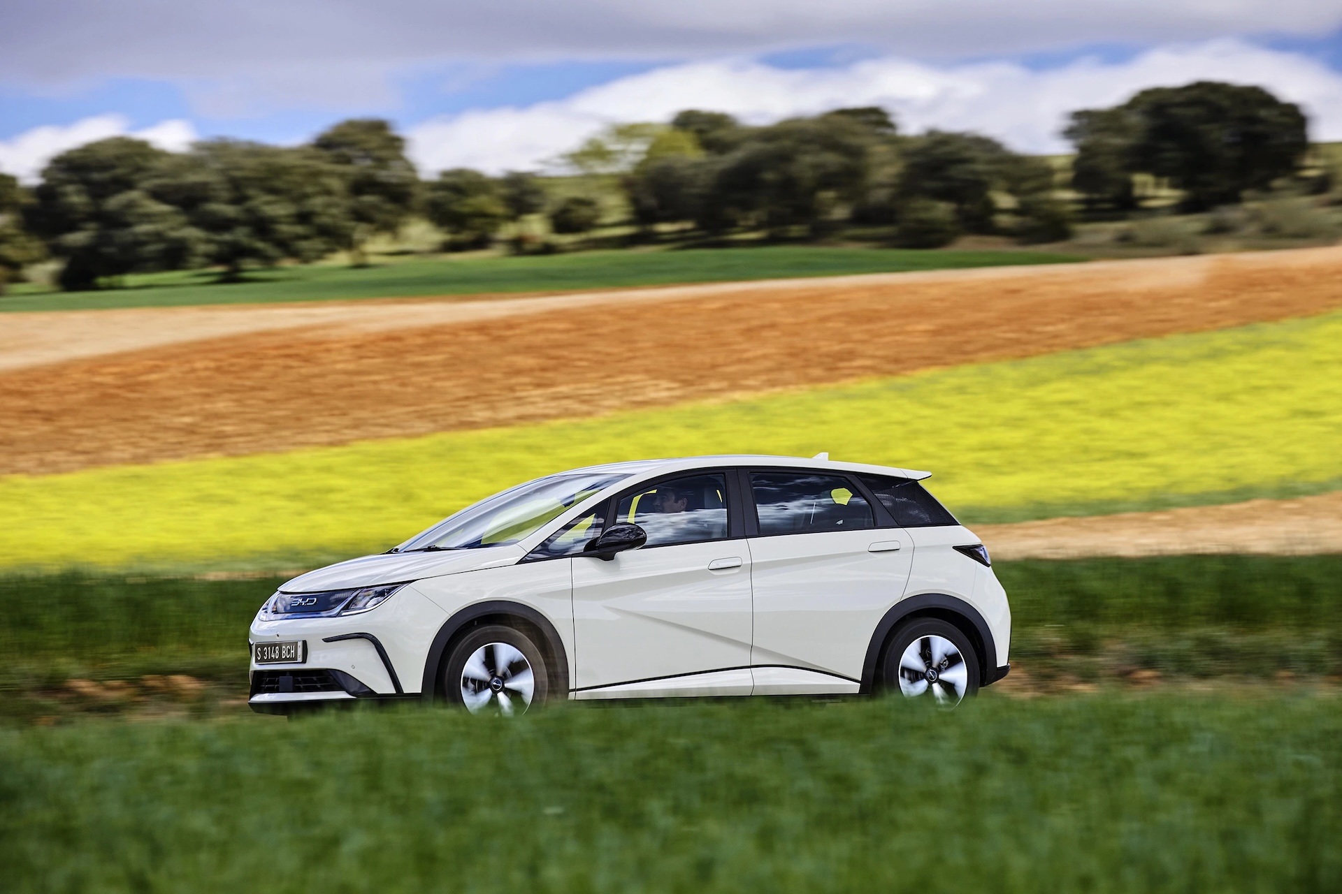 Biały hatchback BYD Dolphin w dynamicznym ruchu na trasie obok kolorowych pól uprawnych.