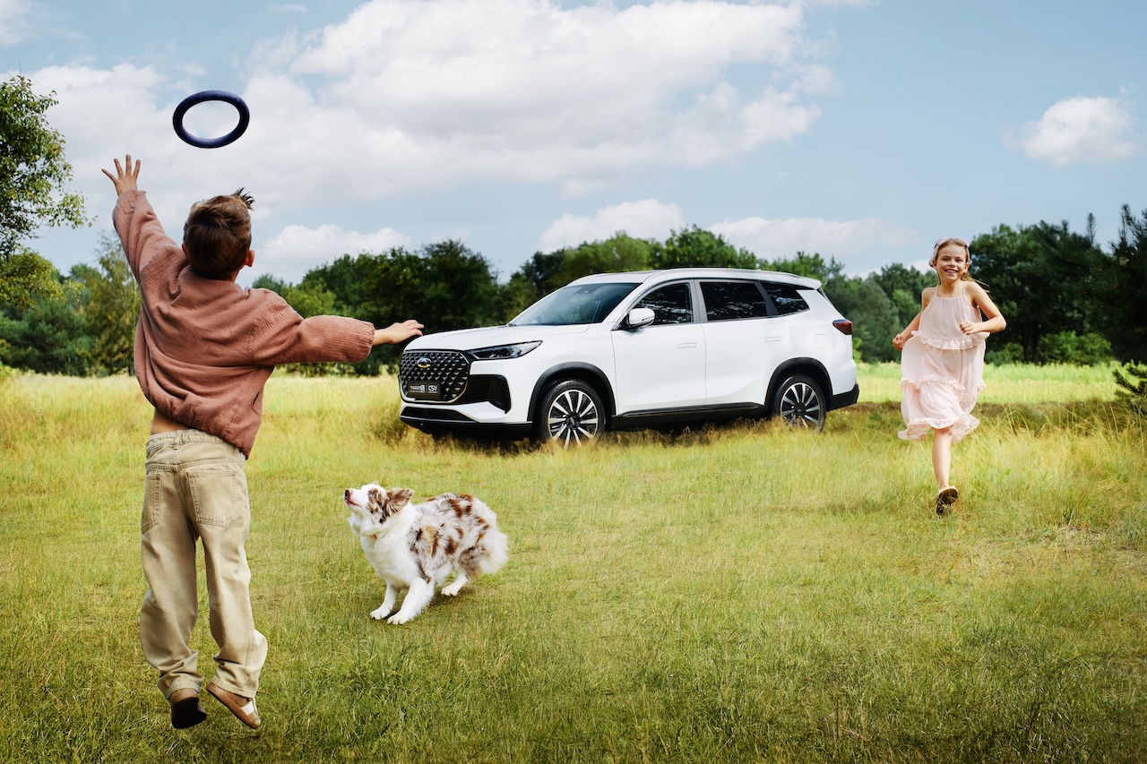 Biały, duży SUV Chery Tiggo 8 stoi na zielonej polanie, w tle bawią się dzieci z psem.