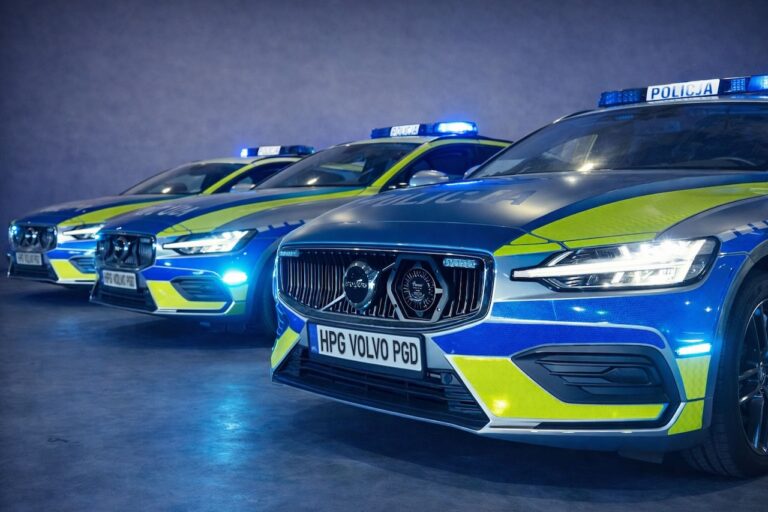Trzy oznakowane radiowozy Volvo V60 w niebiesko-żółtych barwach z włączonymi sygnałami świetlnymi, zaparkowane obok siebie.