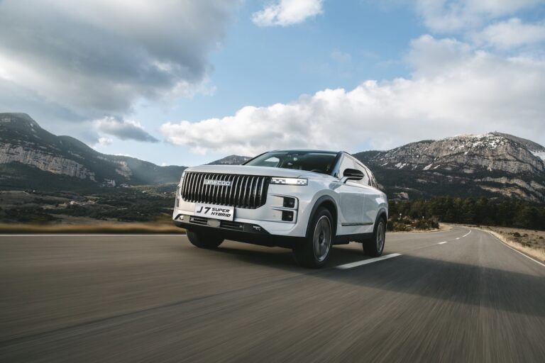 Biały SUV JAECOO 7 jadący po asfaltowej drodze na tle górskiego krajobrazu z zachmurzonym niebem. Na tablicy widnieje napis J7 Super Hybrid.