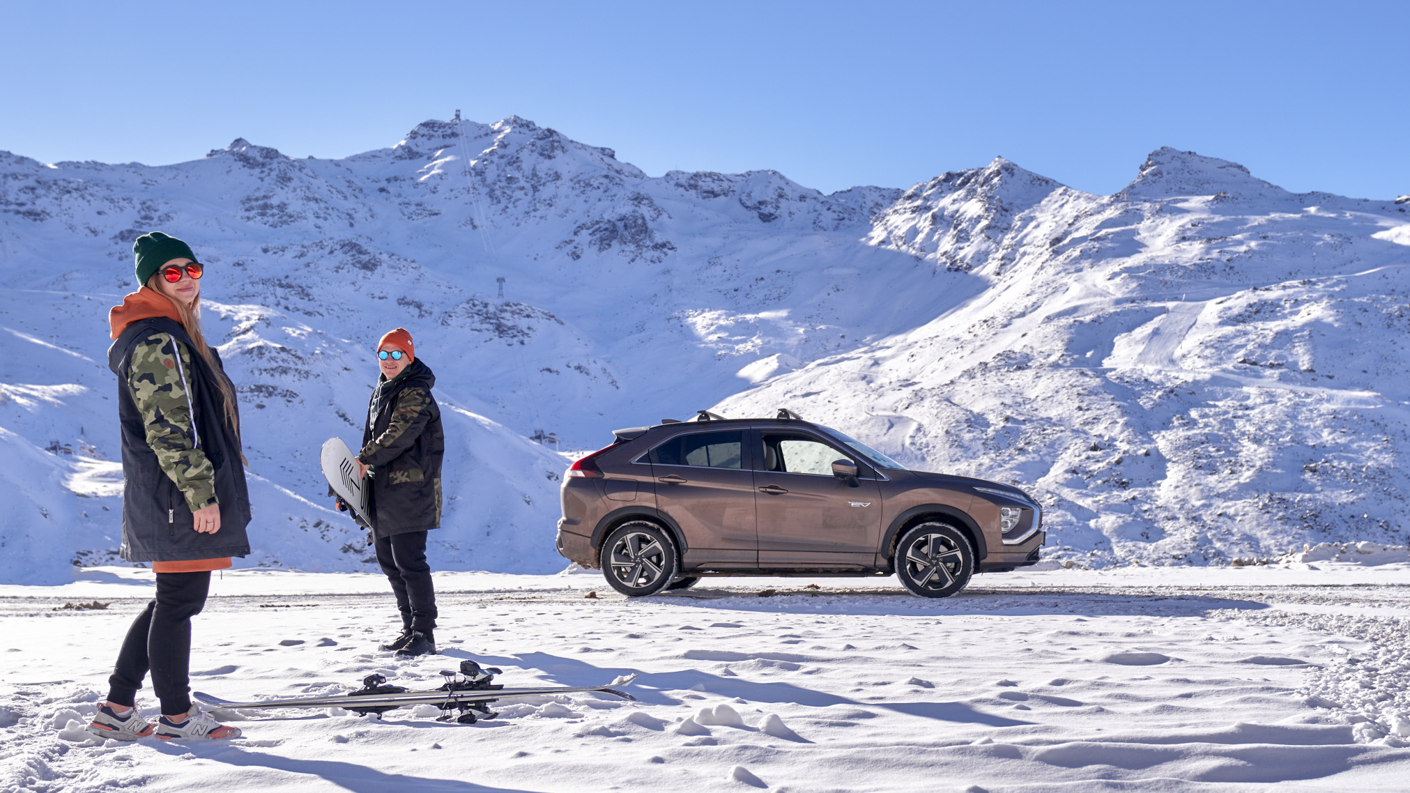 Beżowy samochód typu SUV na tle ośnieżonych górskich szczytów. Para młodych ludzi z deskami snowboardowymi Mitsubishi Eclipse Cross - zimowa promocja Mitsubishi. Lista kontrolna czynności przed zimą