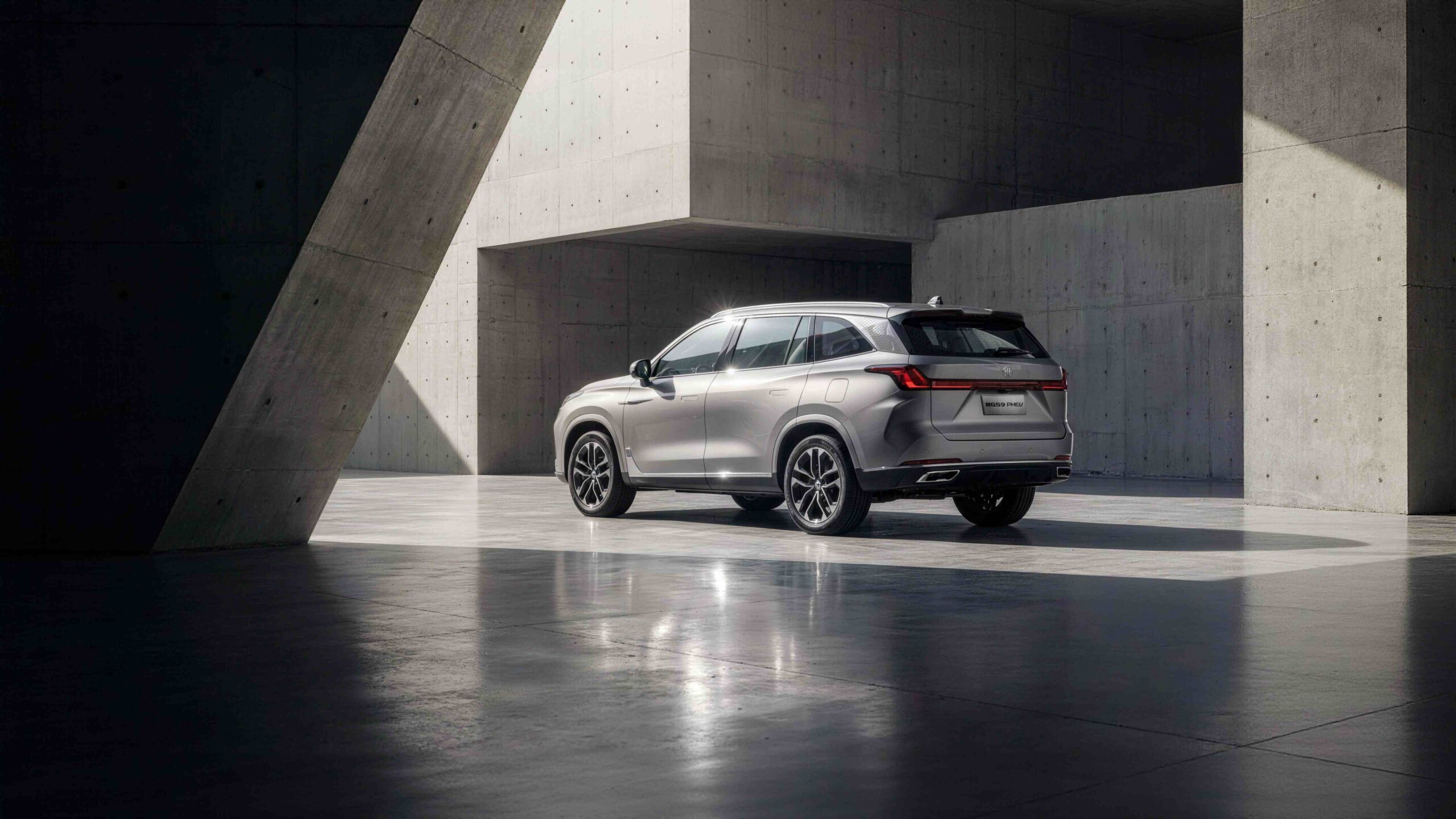 Atrybut Alt: Srebrny samochód MG S9 Plug-In Hybrid zaparkowany w surowym, betonowym otoczeniu nowoczesnej konstrukcji architektonicznej.