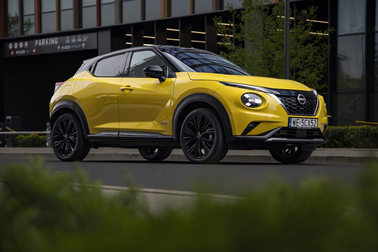 Żółty Nissan Juke z czarnym dachem i czarnymi felgami, zaparkowany przy krawężniku na tle nowoczesnego biurowca z widocznym wjazdem na parking.