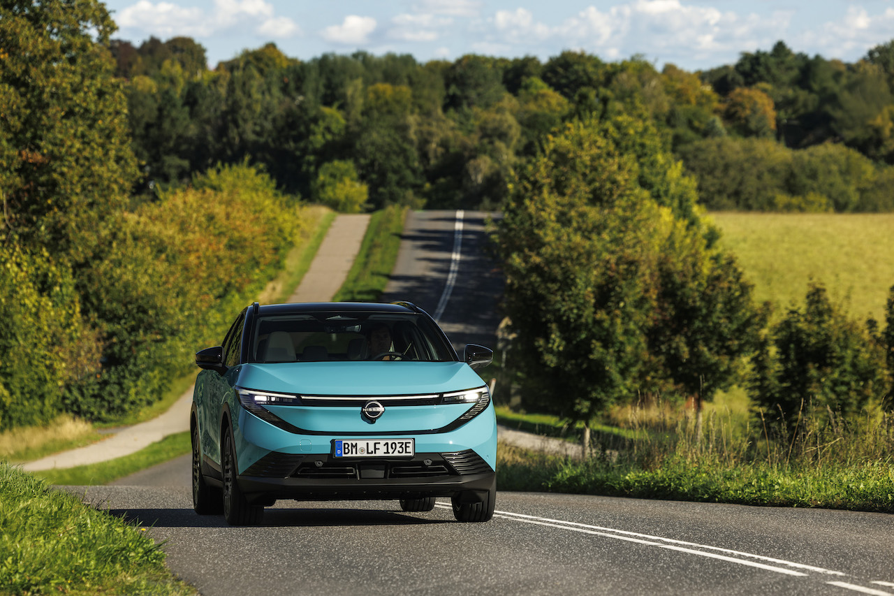 Przód turkusowego Nissana LEAF trzeciej generacji jadącego po asfaltowej drodze w otoczeniu zielonych drzew i wzgórz.