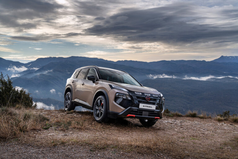 Beżowy Nissan X-Trail w wersji N-Trek zaparkowany na skalistym zboczu na tle rozległych, zamglonych gór i zachmurzonego nieba.