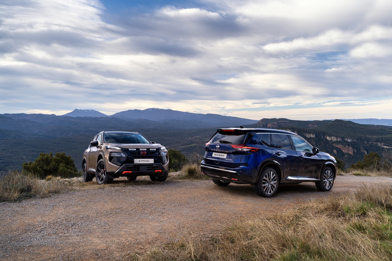 Dwa modele Nissan X-Trail – beżowy oraz granatowy – stojące na szutrowej drodze na tle rozległej panoramy gór pod zachmurzonym niebem.