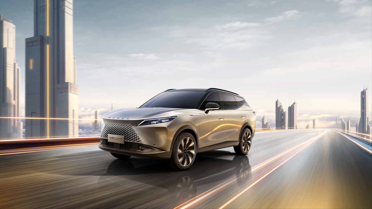 Srebrzysto-złoty SUV OMODA 7 jadący po nowoczesnej trasie na tle futurystycznych wieżowców. Widoczny bezramkowy, geometryczny wlot powietrza oraz wąskie reflektory LED.