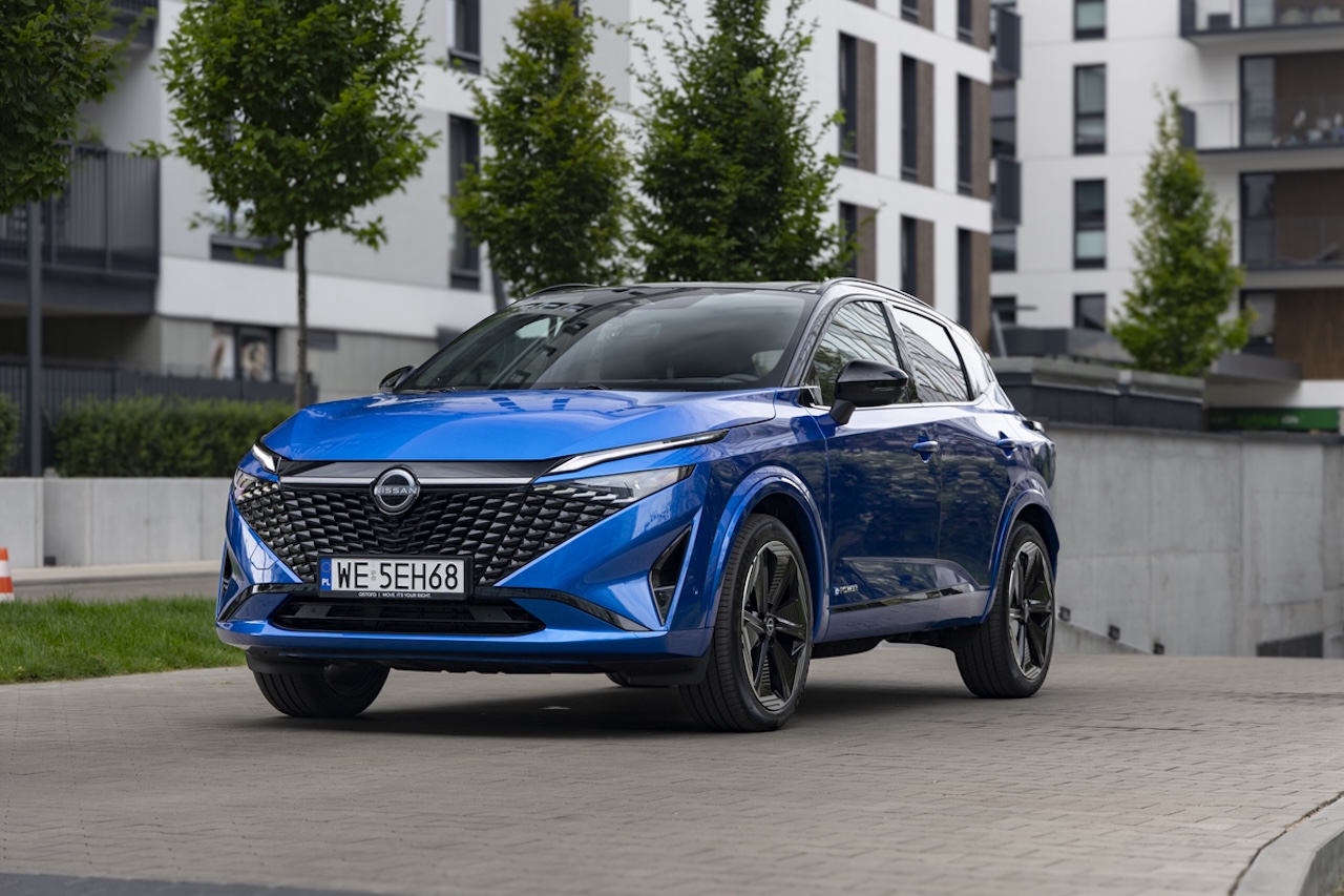 Niebieski Nissan Qashqai zaparkowany na brukowanym podjeździe przed nowoczesnym, białym budynkiem mieszkalnym z zielonymi drzewami w tle.