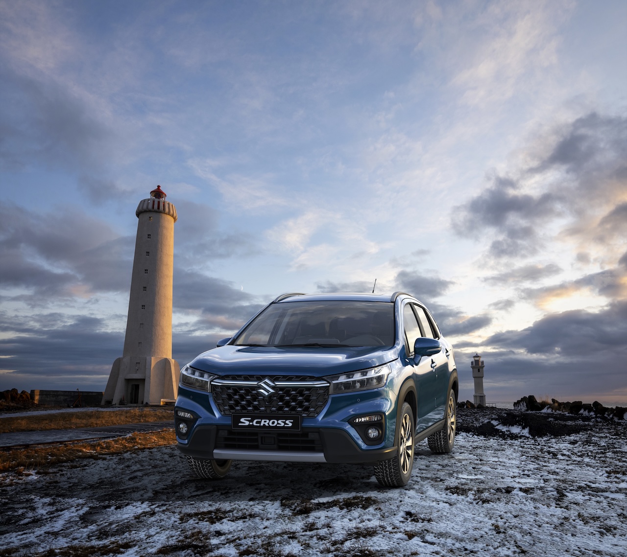 Niebieski SUV Suzuki S-Cross zaparkowany na zaśnieżonym, skalistym brzegu morza obok wysokiej, jasnej latarni morskiej pod zachmurzonym niebem.