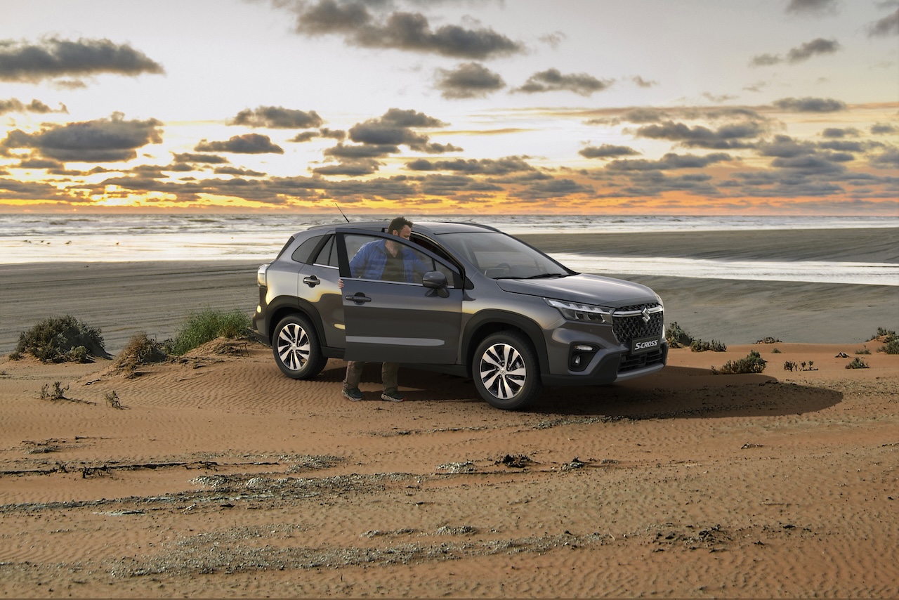 Mężczyzna wsiada do szarego Suzuki S-Cross zaparkowanego na piaszczystych wydmach nad morzem podczas zachodu słońca.