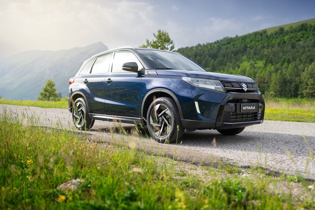 Ciemnoniebieski SUV Suzuki Vitara na asfaltowej drodze w górskim krajobrazie z zielonymi zboczami i słonecznym niebem w tle.
