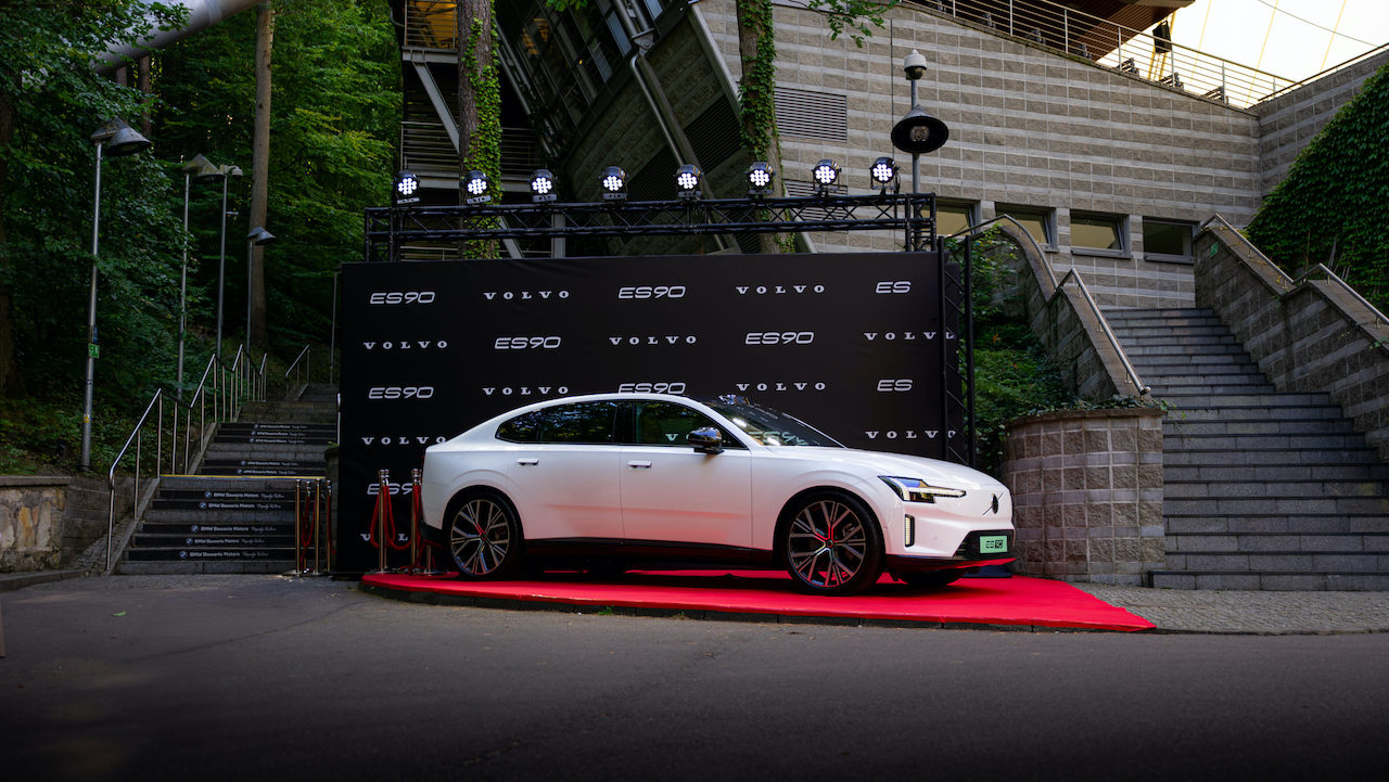 Biały sedan Volvo ES90 zaparkowany na czerwonym podwyższeniu na tle czarnej ścianki z logo marki, umiejscowionej w leśnej scenerii obok schodów.