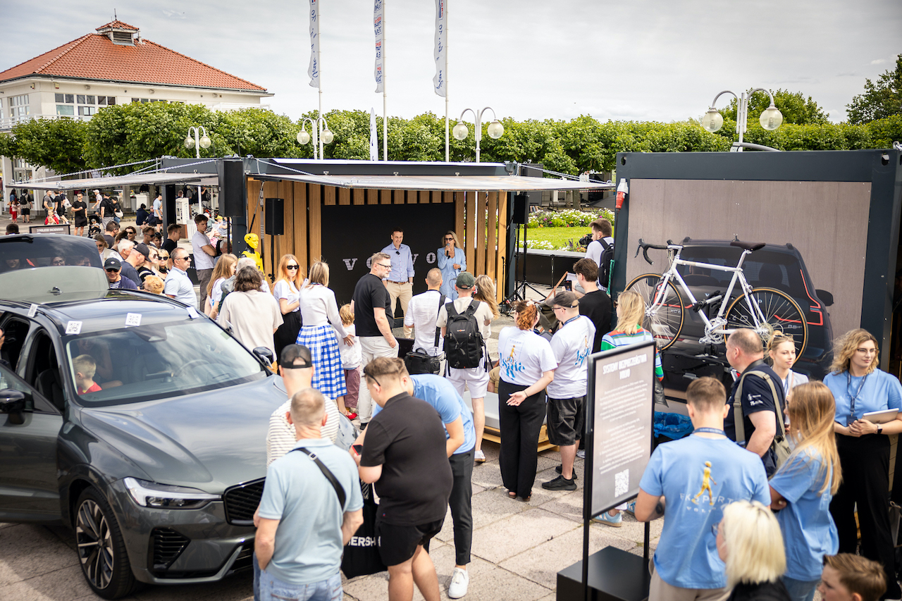 Plenerowe stoisko marki Volvo z kontenerami ekspozycyjnymi i dużym ekranem, otoczone przez tłum ludzi zainteresowanych technologiami bezpieczeństwa.