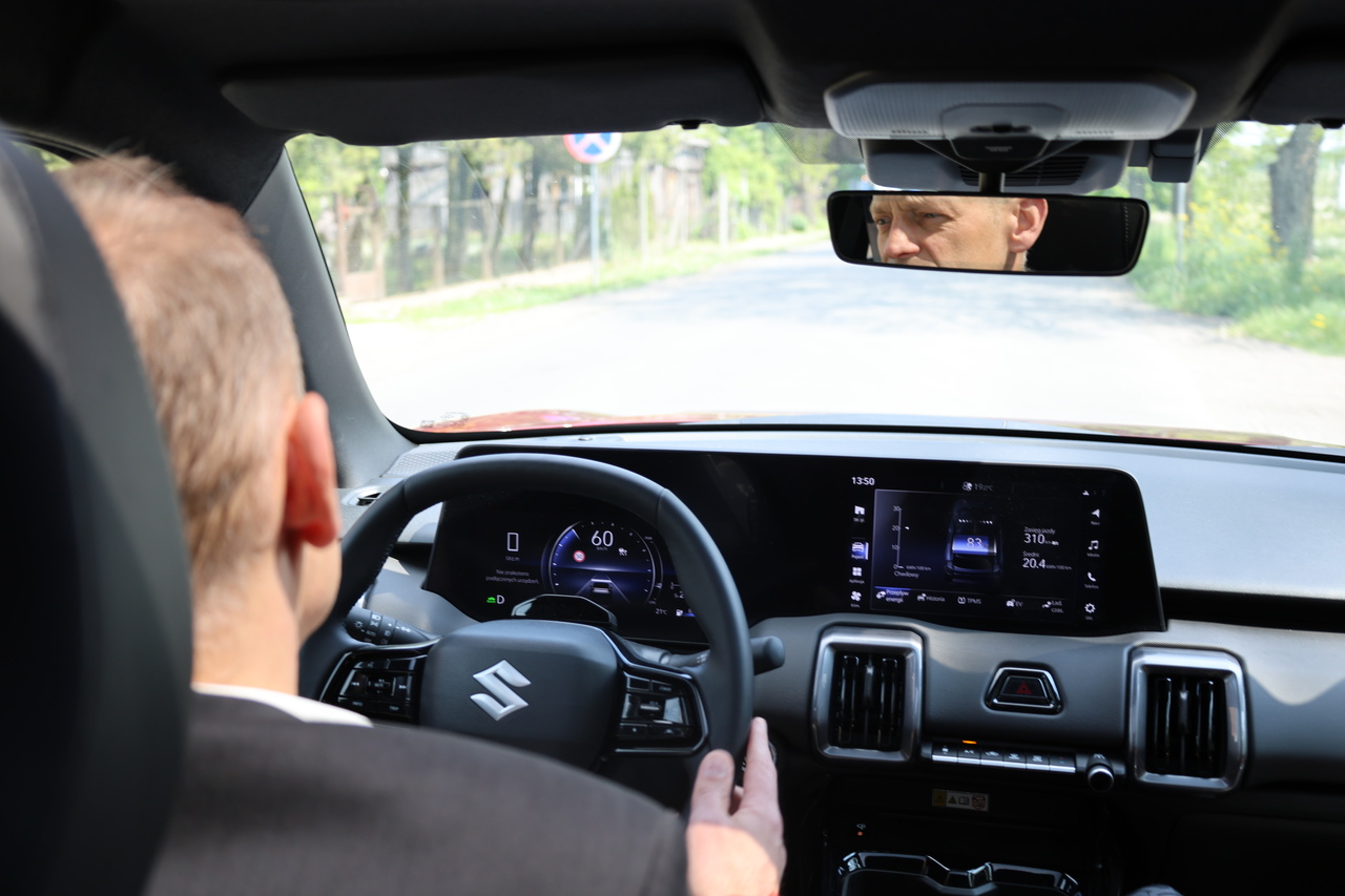 Wnętrze Suzuki e VITARA widoczne zza ramienia kierowcy trzymającego kierownicę, prezentujące nowoczesną deskę rozdzielczą z dwoma dużymi, połączonymi ekranami cyfrowymi pokazującymi parametry jazdy i stan baterii.