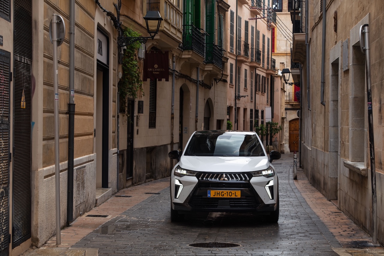 Biały SUV Mitsubishi Grandis zaparkowany przodem na wąskiej, brukowanej uliczce między kamienicami, z wyraźnie widocznym motywem Dynamic Shield na czarnym grillu.