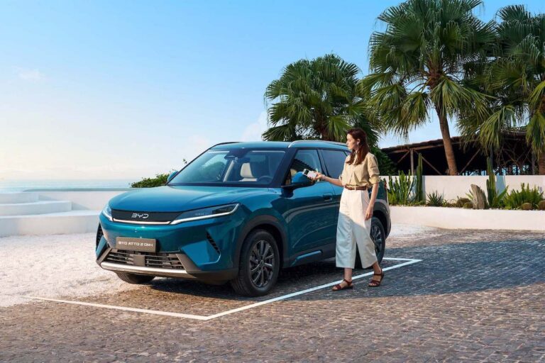 Niebieski SUV BYD ATTO 2 DM-i zaparkowany na brukowanym podjeździe w otoczeniu palm, przy którym kobieta zbliża smartfon do lusterka bocznego, korzystając z funkcji cyfrowego klucza.