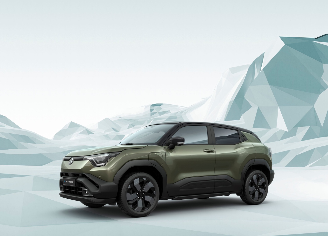 Oliwkowozielony elektryczny SUV Suzuki e VITARA z czarnym dachem, pokazany z profilu na tle jasnego, minimalistycznego krajobrazu z geometrycznymi, trójwymiarowymi górami.
