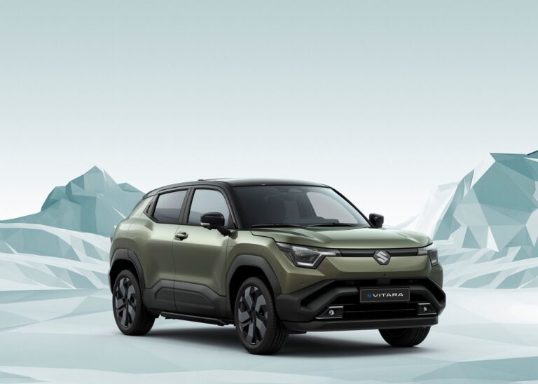 Widok z przodu pod kątem na oliwkowozielony samochód Suzuki e VITARA z wyrazistymi reflektorami LED, dużym logo na grillu i tablicą z nazwą modelu, umieszczony na tle jasnoszarych, wielokątnych brył.