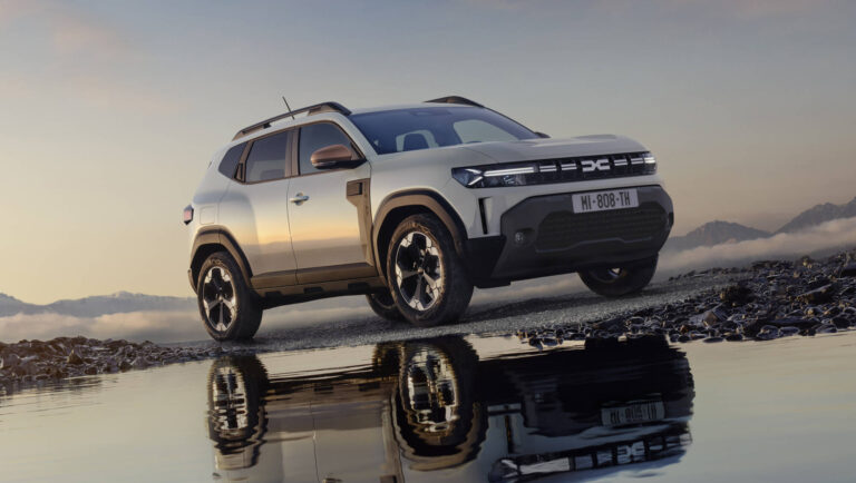 Srebrna Dacia Duster zaparkowana na kamienistym podłożu o zachodzie słońca, z wyraźnym odbiciem sylwetki auta w kałuży na pierwszym planie.