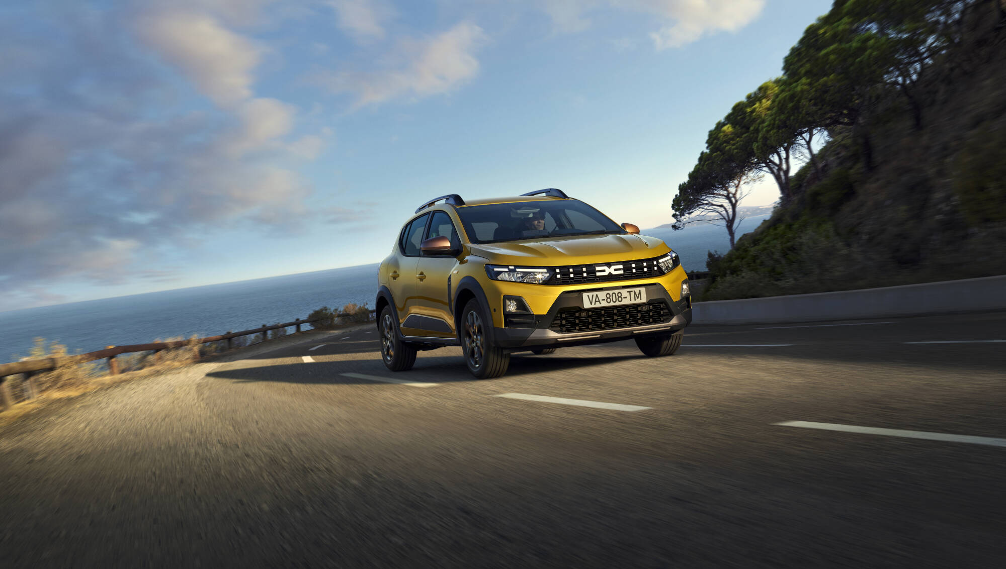 Złota Dacia Sandero Stepway jadąca krętą, nadmorską trasą pod bezchmurnym, błękitnym niebem.