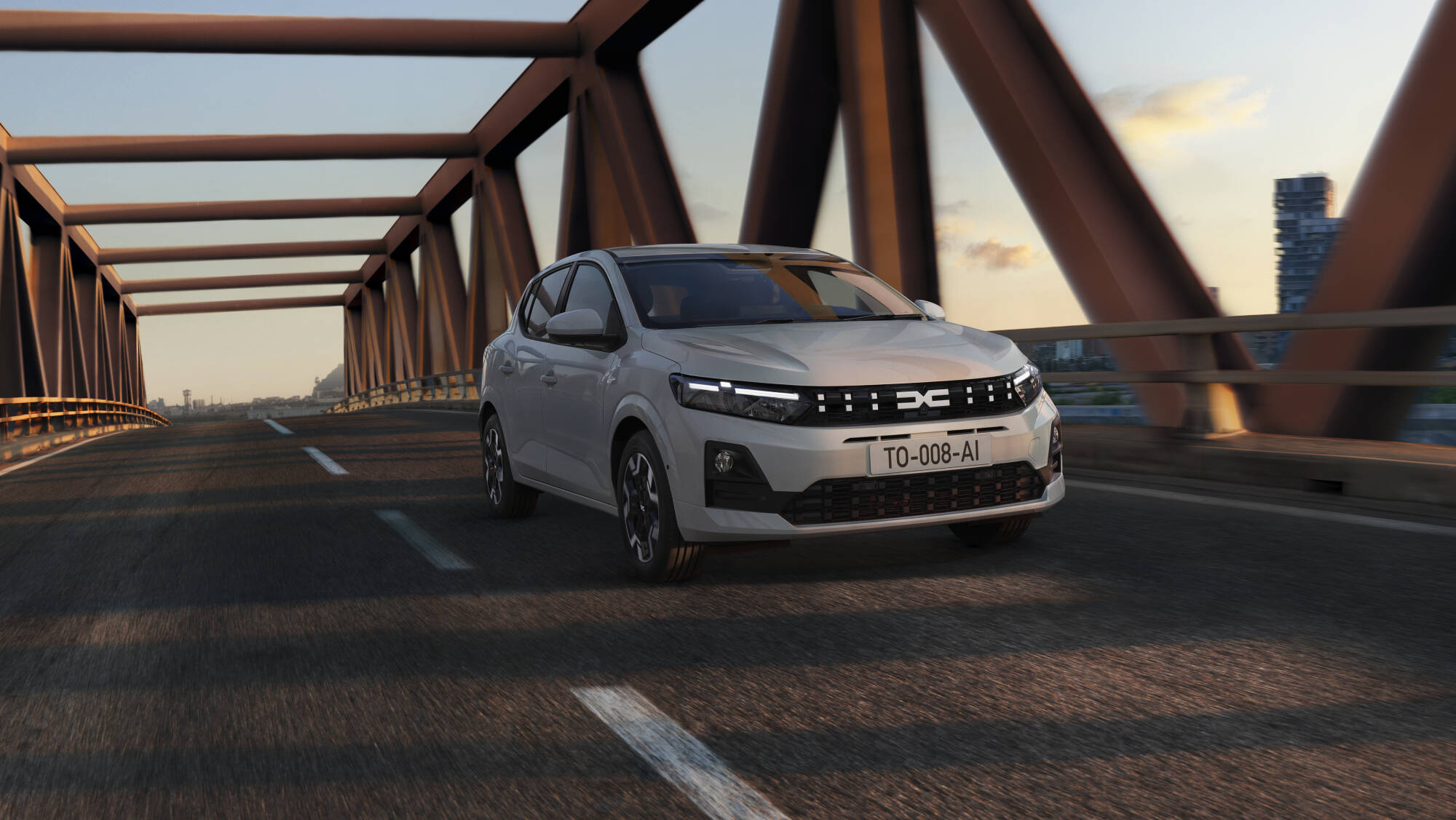 Biała Dacia Sandero przejeżdżająca przez stalowy most o rdzawym kolorze na tle nowoczesnej panoramy miasta.