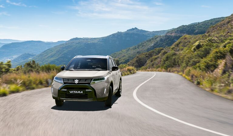 Beżowe Suzuki Vitara jadące krętą asfaltową drogą przez zielone, górzyste tereny pod bezchmurnym niebem.
