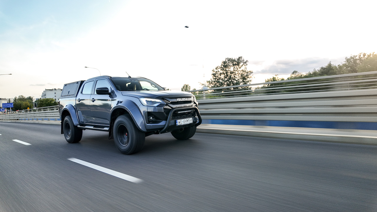 Widok z boku na jadącego po asfalcie ISUZU D-Max Arctic Truck 2025 na tle barier energochłonnych.