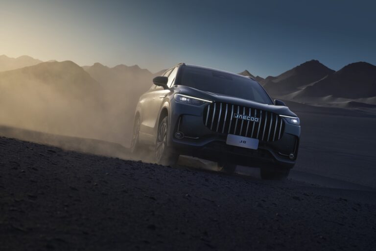 Ciemnoszary SUV JAECOO 8 jadący dynamicznie po piaszczystym wzniesieniu o zmierzchu, z wyraźnie widocznym pionowym grillowaniem i podświetlonymi reflektorami LED.