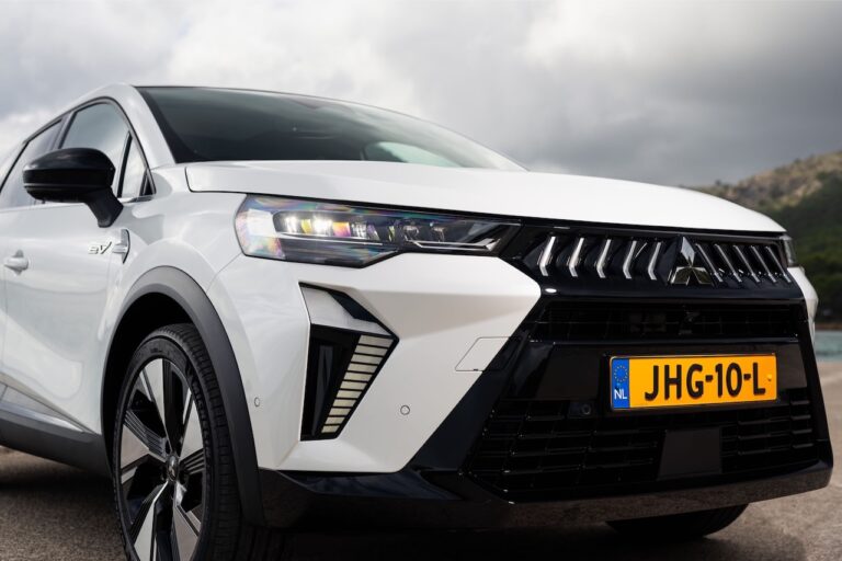 Zbliżenie na przód białego Mitsubishi Grandis z widocznym logo marki, nowoczesnymi reflektorami LED oraz oznaczeniem napędu hybrydowego na przednim błotniku.