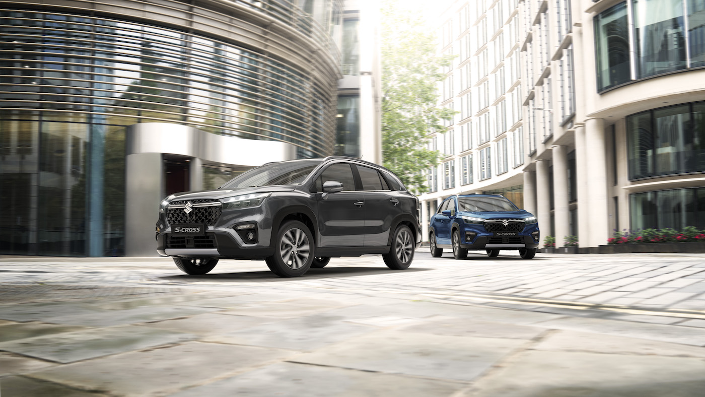 Dwa modele Suzuki S-Cross – ciemnoszary i niebieski – zaparkowane na tle nowoczesnych biurowców