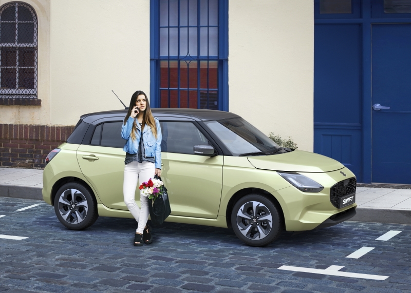 Kobieta z kwiatami obok jasnozielonego Suzuki Swift zaparkowanego przy miejskiej kamienicy