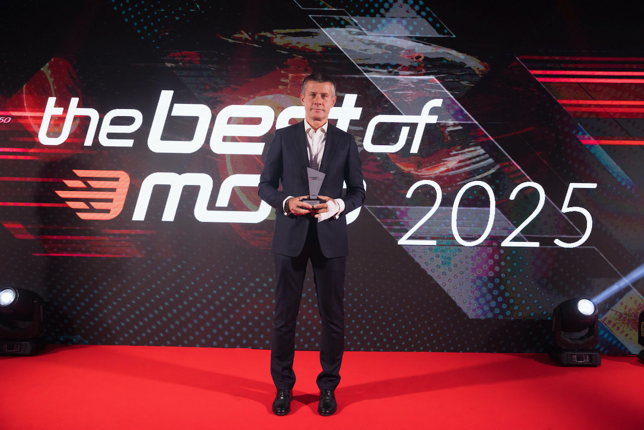 Arkadiusz Nowiński w eleganckim garniturze trzyma statuetkę, stojąc na czerwonym dywanie przed dużym logo plebiscytu „the best of moto 2025”.